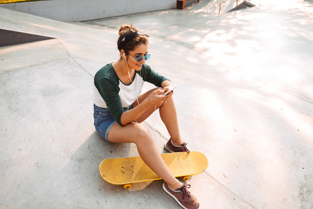 Pretty young girl in earphones listening to music while sitting with skateboard at a park and using mobile phone - Φωτογραφία, εικόνα