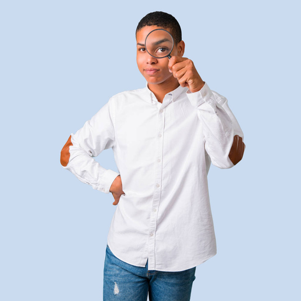 Молодой африканский американец в белой рубашке берет увеличительное стекло и смотрит сквозь него на изолированном голубом фоне
 - Фото, изображение
