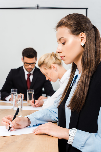 attractive young businesswoman writing on blank paper during conference with colleagues - Photo, Image