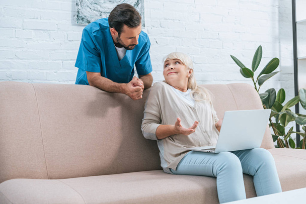 nuori mies sairaanhoitaja nojaa sohvalla ja katselee vanhempi nainen käyttää kannettavaa tietokonetta
  - Valokuva, kuva