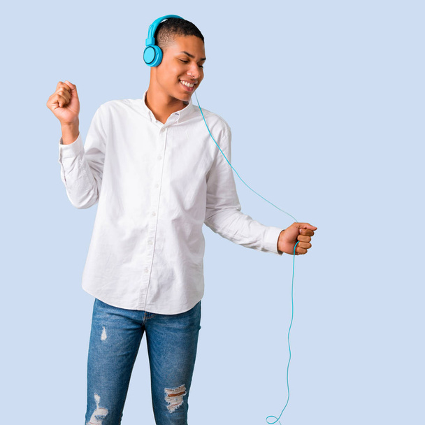 Young african american man with white shirt listening to music with headphones and dancing on isolated blue background - Photo, Image