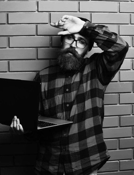 hipster holding laptop in red black checkered shirt with hat and glasses on brown brick wall studio background. Bearded brutal caucasian hipster holding laptop - Foto, afbeelding
