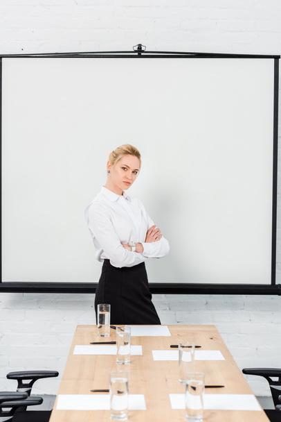 hermosa joven mujer de negocios con brazos cruzados delante del tablero de presentación en blanco en la sala de conferencias
 - Foto, imagen