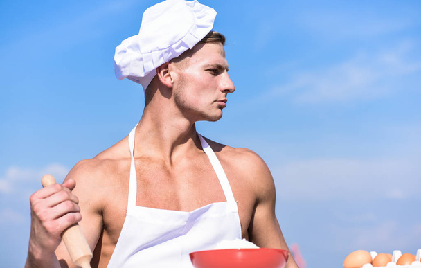 Man on confident face wears cooking hat and apron, sky on background. Chef cook preparing dough for baking. Cook or chef with sexy muscular shoulders and chest holds rolling pin. Cookery concept - Foto, Imagen