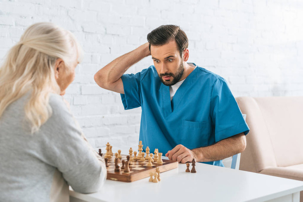 confused male caregiver playing chess with senior woman - Zdjęcie, obraz