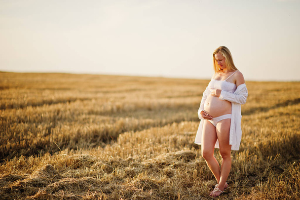 Blonde pregnant woman in wreath field at white underwear clothes on sunset. Happy moments of pregnancy. - Foto, immagini