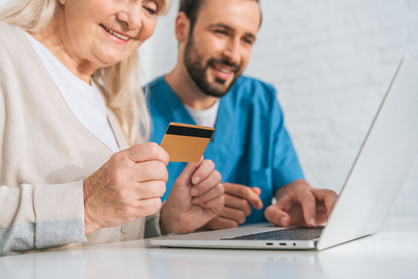przycięte strzał uśmiechający się starszy kobieta posiadania karty kredytowej i za pomocą laptopa z młodego pracownika socjalnego - Zdjęcie, obraz