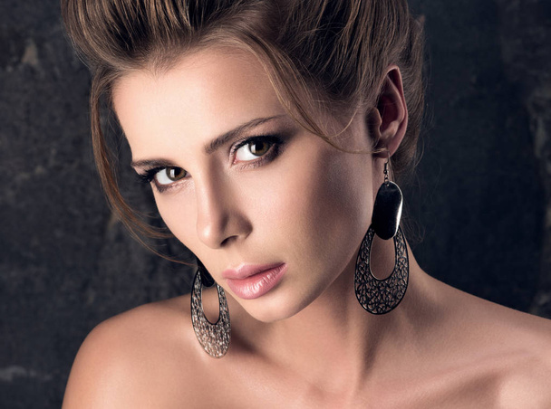 Beautiful young woman with sensual look. Professional makeup, hairstyle and big earrings. The dark background - Photo, Image