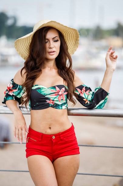 Outdoor portrait of beauty brunette model in colorful summer clothes. Young beautiful woman standing on embankment at daytime, blurred background  - Фото, зображення