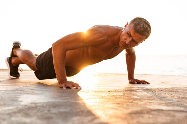 Confident sportsman doing push-ups at the beach in sunlight - Fotó, kép