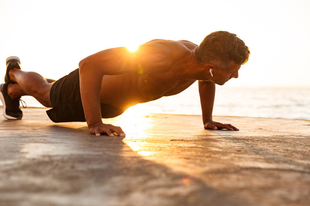 Confident sportsman with earphones doing push-ups at the beach in sunlight - Photo, Image