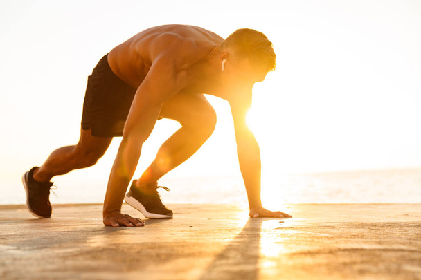 Confident sportsman with earphones getting ready to run at the beach in sunlight - Foto, afbeelding
