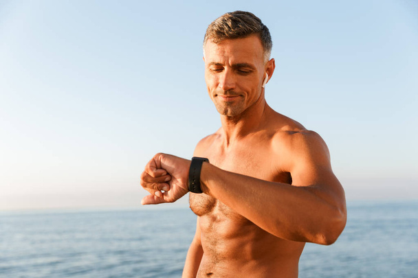 Close up of smiling handsome shirtless sportsman with earphones checking his smart watch at the beach - Foto, immagini