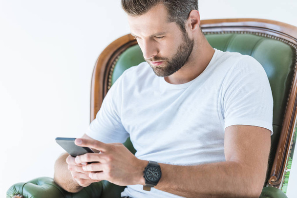 handsome bearded man using smartphone while sitting in armchair, isolated on white - Photo, image