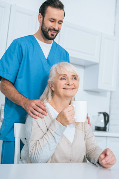Lächelnder männlicher Pfleger schaut Seniorin beim Teetrinken in Küche an  - Foto, Bild