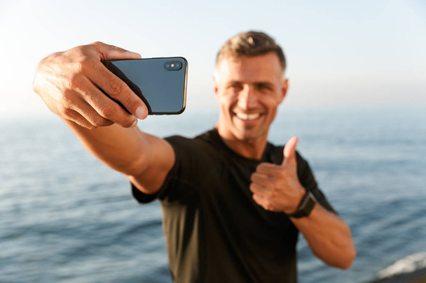 Smiling handsome shirtless sportsman taking a selfie while standing at the beach and showing thumbs up - Foto, imagen