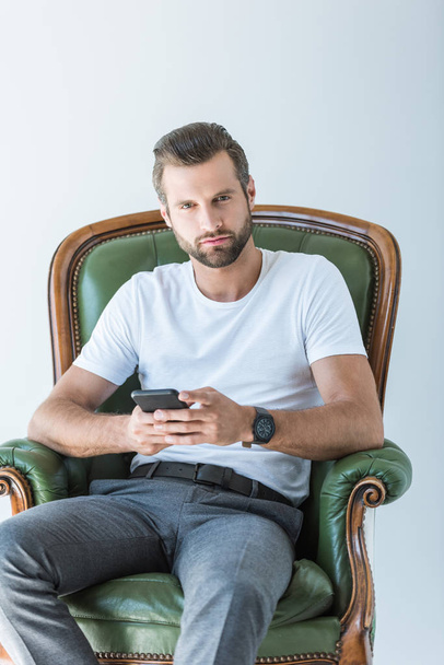 thoughtful bearded man using smartphone while sitting in armchair, isolated on white - Foto, Imagem