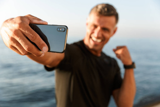 Confident handsome shirtless sportsman taking a selfie while standing at the beach and flexing biceps - 写真・画像