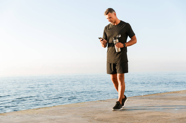 Full length of smiling sportsman using mobile phone while standing at the beach and holding bottle of water - Photo, image