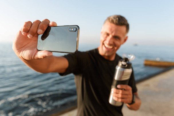 Image of handsome mature sportsman take a selfie with bottle of water on the beach. - Photo, image
