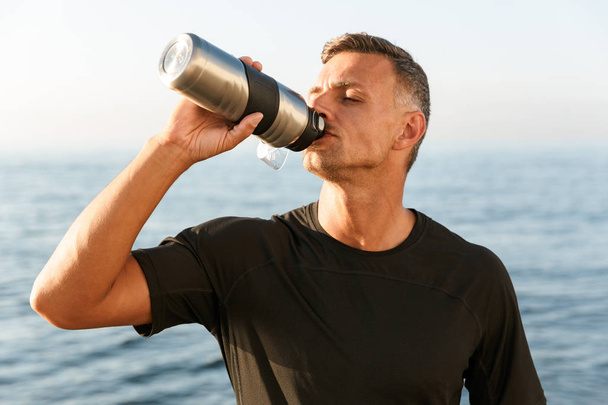 Image of strong mature sportsman standing with bottle of water drinking on the beach. - Foto, afbeelding