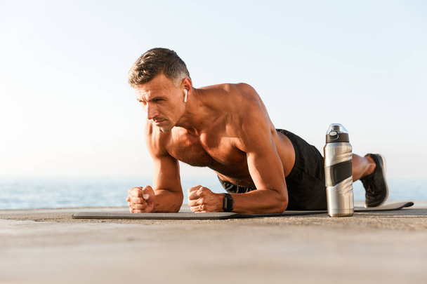 Concentrated shortless sportsman in earphones doing plank exercises on a fitness mat at the beach - Фото, изображение