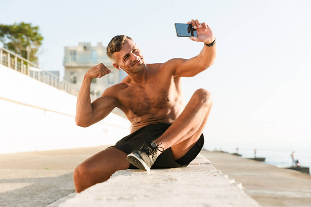 Esportista sem camisa bonito sorridente com fones de ouvido tirando uma selfie e bíceps flexionando enquanto sentado na praia
 - Foto, Imagem