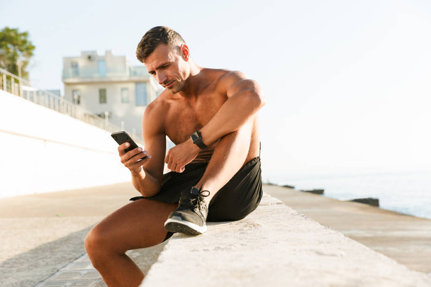 Photo of strong mature sportsman chatting by mobile phone on the beach outdoors. - Foto, Bild