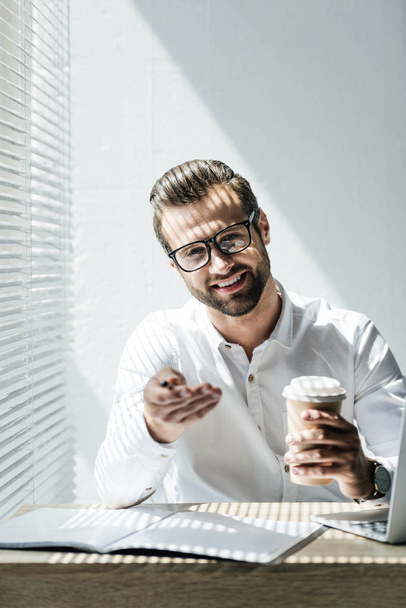 χαμογελώντας επιχειρηματία σε γυαλιά, κάθεται στο χώρο εργασίας με φορητό υπολογιστή και γραφειοκρατία στο διάλειμμα για καφέ - Φωτογραφία, εικόνα