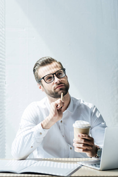 pensativo hombre de negocios sentado en el lugar de trabajo con el ordenador portátil, papeleo y taza de café desechable
  - Foto, imagen