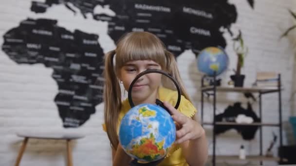 Portrait of a cute girl who playing looking in magnifying glass. Girl looking through a magnifying glass and studies the globe or map of the world - Metraje, vídeo