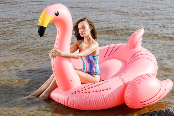 Outdoor portrait of beauty brunette model in colorful summer swimwear. Young beautiful woman sitting on pink inflatable mattress in form of flamingo at daytime, wavy water background - 写真・画像
