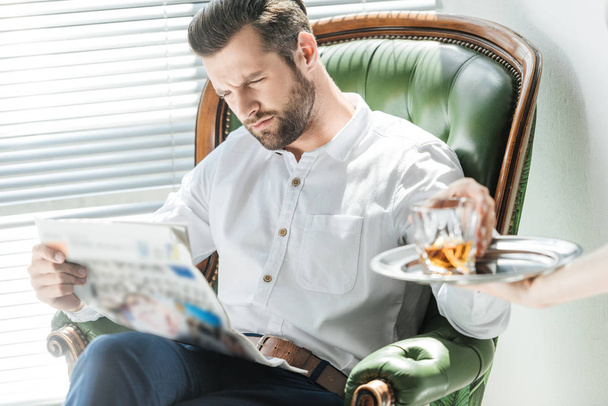 hombre serio con estilo leyendo periódico mientras toma vaso de whisky de la bandeja
 - Foto, Imagen