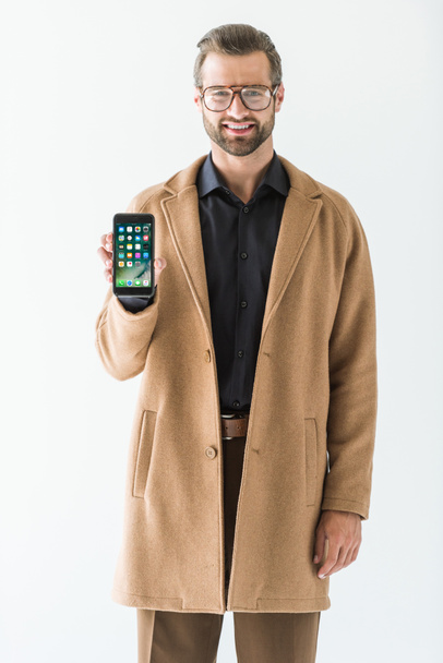 бородатый улыбающийся мужчина в пальто представляет iphone, изолированный на белом
 - Фото, изображение