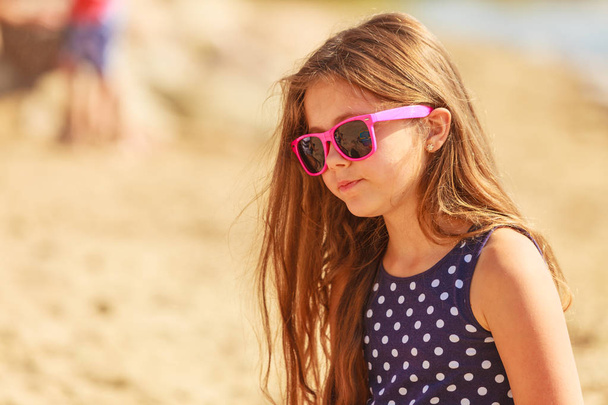 Летние каникулы и отдых. Маленькая девочка-туристка в розовых солнечных очках на улице. Ребенок ждет родителя на пляже
. - Фото, изображение