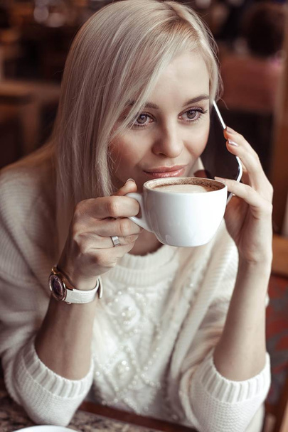 Portrait of a beautiful young blonde. A happy young woman in a knitted sweater is sitting in a cafe drinking cappuccino and talking on the phone. Autumn. Winter. Cozy. - Photo, Image