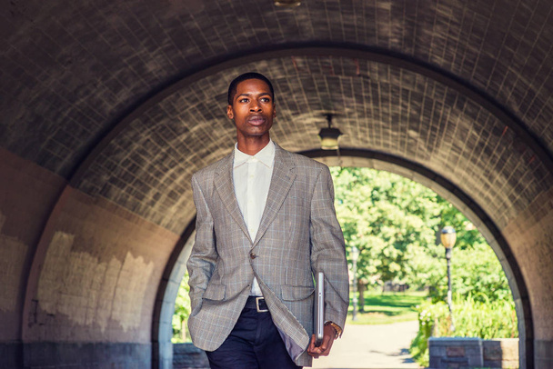 Νέοι Αφρικανικός Αμερικανός επιχειρηματίας ταξιδεύει στη Νέα Υόρκη φορώντας μοτίβο blazer, λευκό εσώρουχο, μαύρο παντελόνι, κρατώντας το φορητό υπολογιστή, περπατώντας κάτω από την γέφυρα του δρόμου, με σιγουριά με ανυπομονησία. - Φωτογραφία, εικόνα