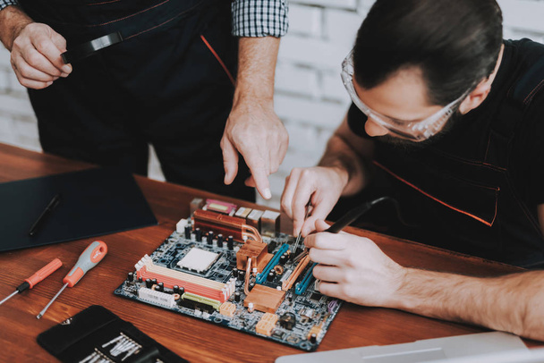 Two Men Repairing Hardware Equipment in Workshop. Repair Shop. Worker with Tools. Computer Hardware. Young and Old Workers. Modern Devices. Digital Device. Laptop on Desk. Electronic Devices Concept. - 写真・画像
