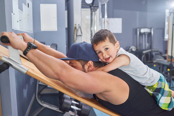 Young father with strong muscles training in gym keeping little son on his back. - Фото, изображение