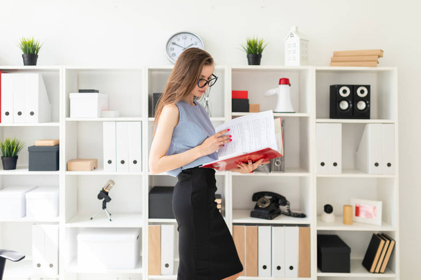 Mooi jong meisje in een kantoor staat in de buurt van een rack en bladert door een map met documenten. - Foto, afbeelding