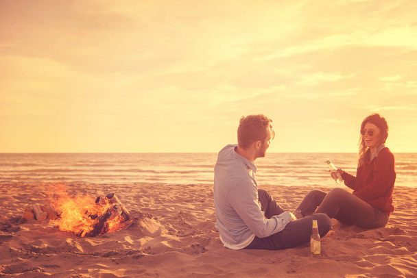 Young Couple Relaxing By The Fire, Drinking A Beer Or A Drink From The Bottle on the beach at autumn day filter - Photo, image