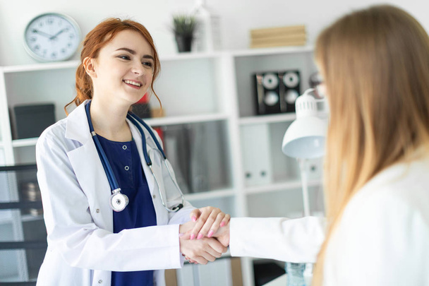 A beautiful young girl in a white coat is standing near the desk in the office and shaking hands with the patient. The patient thanks the doctor. - Photo, image