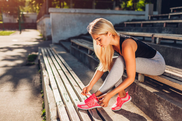 Jonge vrouw atleet koppelverkoop sneakers zittend op de Bank op sportsground in de zomer. Voorbereiding op de opleiding. Training - Foto, afbeelding