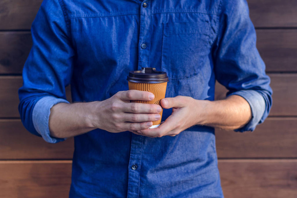 Człowiek w jeans koszula trzymając kubek świeżej kawy na brązowym tle - Zdjęcie, obraz