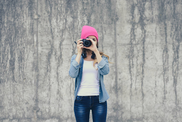 Hipster fille en jeans vêtements et chapeau rose prenant des photos contre le mur de béton gris. Focus sur la caméra
 - Photo, image