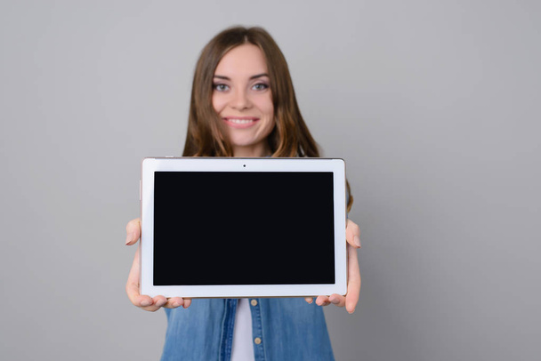 Atrakcyjna kobieta z Uśmiech zębiczny Wyświetlono cyfrowego tabletu z czarny pusty ekran dotykowy. Kobieta jest na białym tle na szarym tle - Zdjęcie, obraz