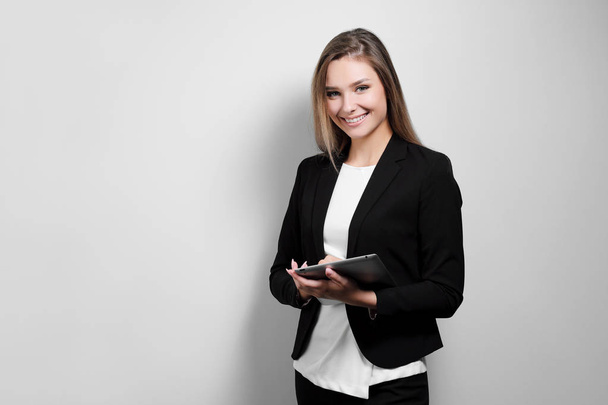 Portrait of a beautiful smiling woman blonde with a tablet in her hands in a business suit and jacket on a white background. A student or a businesswoman is working. - Photo, Image