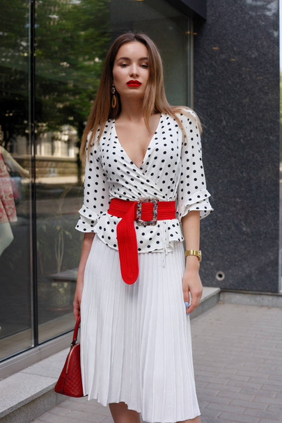 beautiful model look brunette female wearing white dress with black polka dots is while waking on a city street background with stylish bags in the hand. - Fotografie, Obrázek