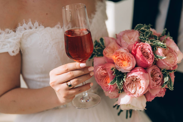 Крупный план обрезанной рамы невесты в белом платье держит в руке свадебный букет розовых пионов и бокал розового шампанского, носит плоское обручальное золотое кольцо. Маникюр
 - Фото, изображение