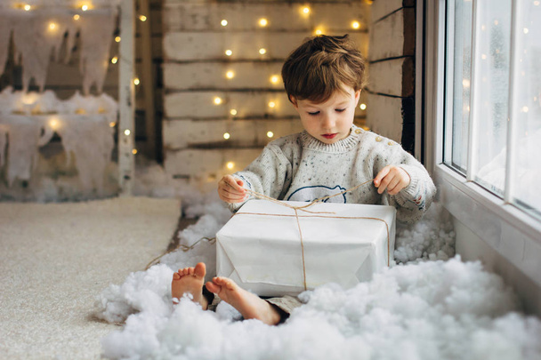 Beaming curlyhair boy opening his christmas gift. New Year lights background - Φωτογραφία, εικόνα